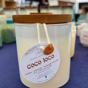 Puede incluir: Una vela cil&iacute;ndrica en un recipiente met&aacute;lico con tapa de madera. La vela tiene una etiqueta que dice "Coco Loco" y enumera los ingredientes. La vela tiene un peso neto de 241 g.