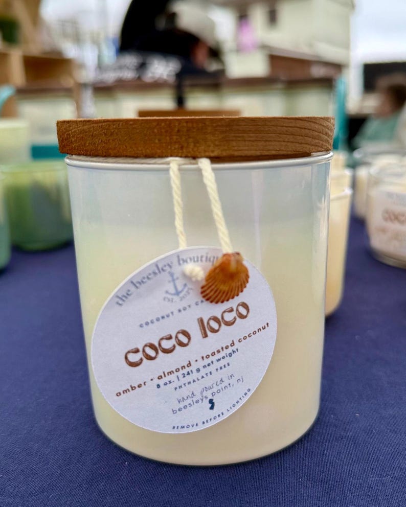 Puede incluir: Una vela cil&iacute;ndrica en un recipiente met&aacute;lico con tapa de madera. La vela tiene una etiqueta que dice "Coco Loco" y enumera los ingredientes. La vela tiene un peso neto de 241 g.