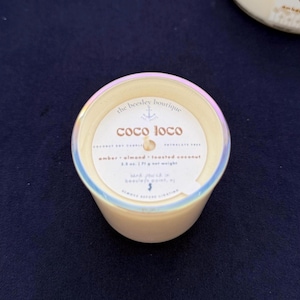 Puede incluir: Una vela peque&ntilde;a y redonda en un recipiente color crema con un borde hologr&aacute;fico. La etiqueta dice "coco loco" y enumera la fragancia como &aacute;mbar, almendra y coco tostado. El peso neto es de 71 g.
