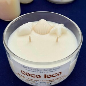 Puede incluir: Un portavelas de cristal transparente lleno de cera blanca cremosa. La vela est&aacute; decorada con adornos de cera con forma de concha. La etiqueta dice "Coco Loco" y enumera los ingredientes. La vela tiene dos mechas.