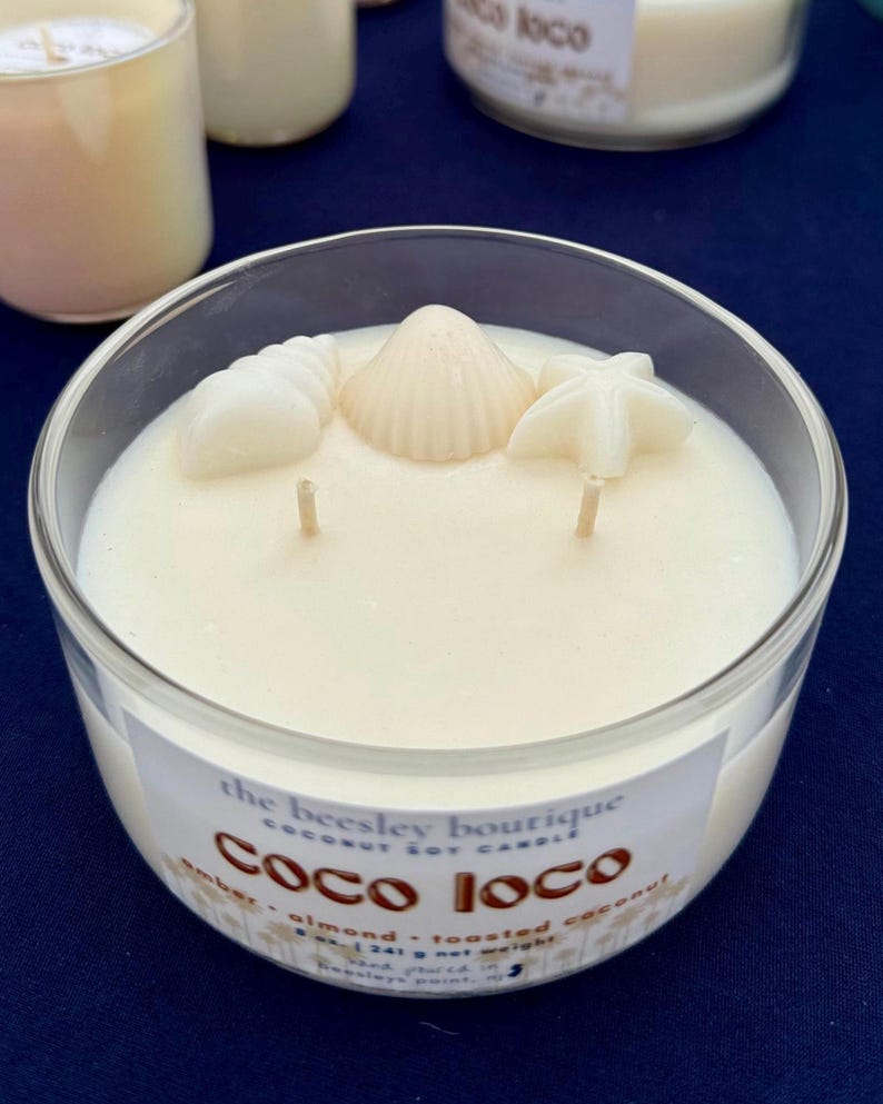 Puede incluir: Un portavelas de cristal transparente lleno de cera blanca cremosa. La vela est&aacute; decorada con adornos de cera con forma de concha. La etiqueta dice "Coco Loco" y enumera los ingredientes. La vela tiene dos mechas.