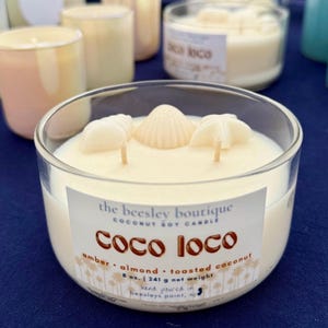 Puede incluir: Un portavelas de cristal transparente lleno de cera cremosa, adornado con tres decoraciones de conchas marinas. La vela tiene tres mechas y una etiqueta que dice "Coco Loco" con notas de aroma. Pesa 241 g y est&aacute; vertida a mano.