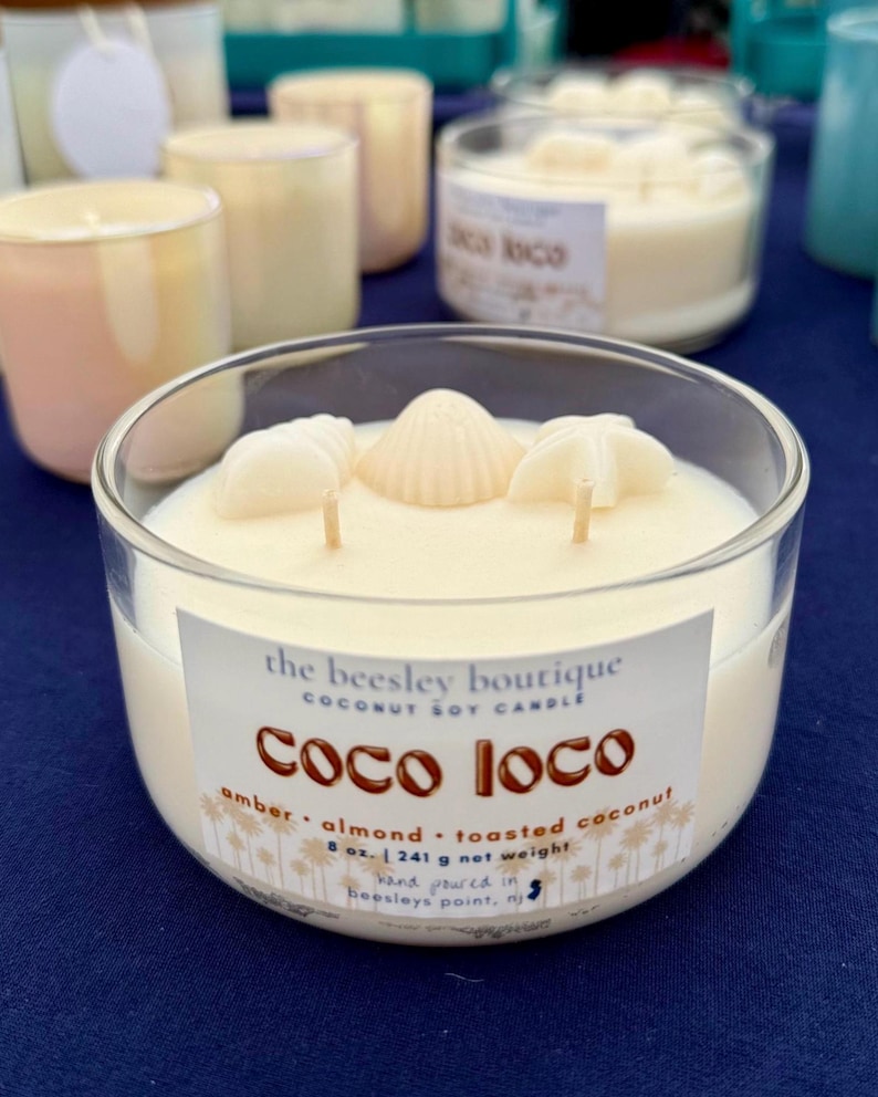 Puede incluir: Un portavelas de cristal transparente lleno de cera cremosa, adornado con tres decoraciones de conchas marinas. La vela tiene tres mechas y una etiqueta que dice "Coco Loco" con notas de aroma. Pesa 241 g y est&aacute; vertida a mano.