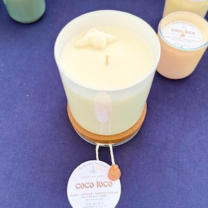 Puede incluir: Una vela blanca en un recipiente de vidrio con un adorno de concha marina, sobre un posavasos de madera. La vela tiene una etiqueta que dice "coco loco" y est&aacute; perfumada con &aacute;mbar, almendras y coco tostado. Otras velas son visibles en el fondo.