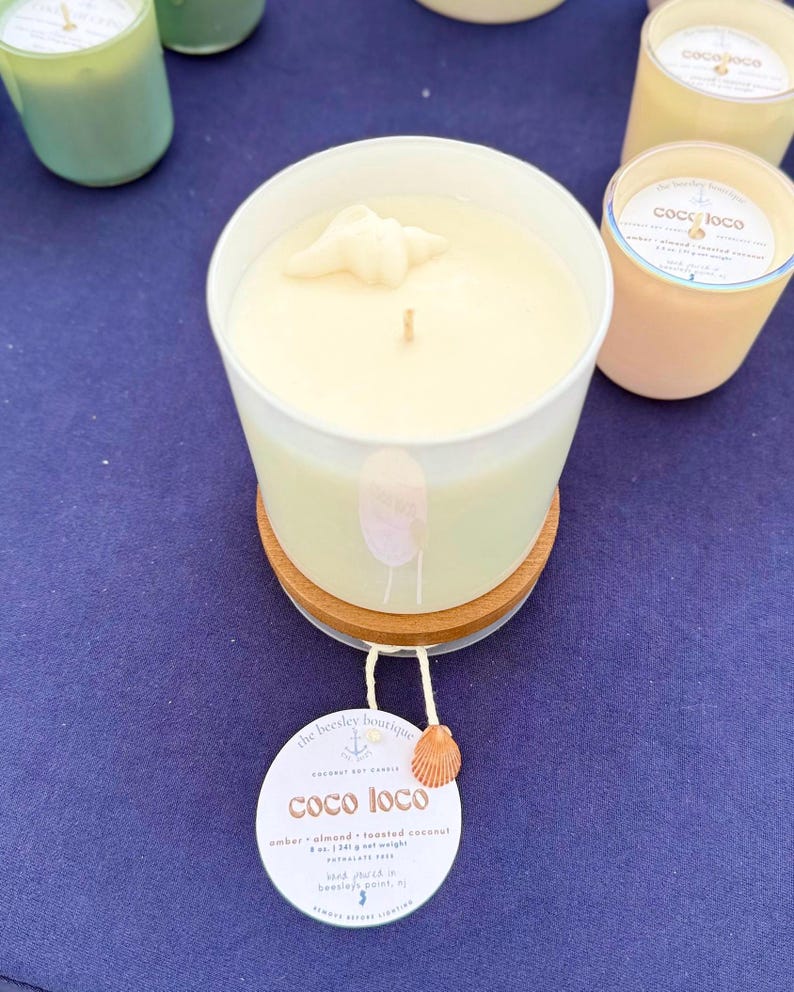 Puede incluir: Una vela blanca en un recipiente de vidrio con un adorno de concha marina, sobre un posavasos de madera. La vela tiene una etiqueta que dice "coco loco" y est&aacute; perfumada con &aacute;mbar, almendras y coco tostado. Otras velas son visibles en el fondo.