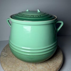 May include: A mint green Le Creuset stoneware crock with a matching lid. The container features two side handles, a rounded body with horizontal ribbed detailing, and the embossed text LE CREUSET on the top of the lid. The finish is glossy.