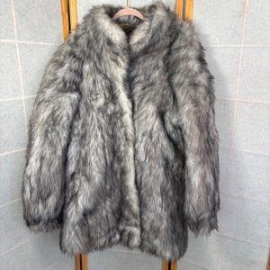 May include: A long, faux fur coat in shades of gray and white hangs on a wooden hanger. The coat has a wide collar and a soft, textured appearance. The fur appears thick and plush, creating a luxurious look.
