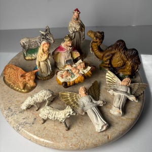 May include: A collection of painted ceramic nativity figurines including a central infant in a manger, Mary, Joseph, a king, a camel, a donkey, an ox, two sheep, and two angels with gold-painted wings. The figures are arranged on a round stone base.