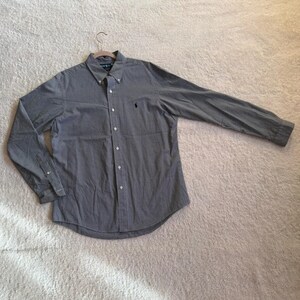 May include: A long-sleeve button-down shirt in a dark navy and white micro-check pattern. The shirt features a pointed collar, a front button placket, and a small black embroidered logo on the left chest. It is displayed on a hanger.