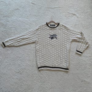 May include: A long-sleeve crewneck sweater in a cream-colored textured knit. The garment features dark brown trim at the collar, cuffs, and hem. Centered on the chest is an embroidered graphic depicting a fishing net and rod.