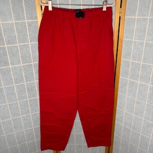 May include: Bright red pants with a black buckle closure at the waist. The pants are made of a solid, opaque material and appear to be a casual style. The pants are hanging on a white hanger.