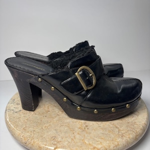 May include: Black leather clogs with a wooden platform and heel. The clogs feature a buckle detail with gold-tone hardware and are lined with black faux fur. The platform is studded with small gold-tone rivets.