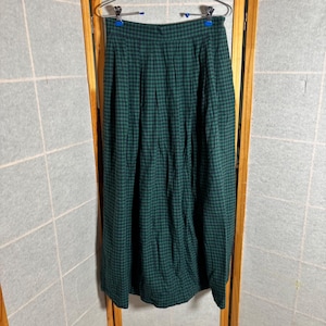 May include: A long, green and black checkered skirt hangs on a hanger. The skirt is made of a soft fabric and has a high waist. The checkered pattern is a classic design.