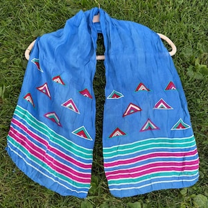 May include: A bright blue scarf featuring a pattern of small, multi-colored triangles in red, white, and teal. The bottom section of the scarf displays horizontal stripes in alternating shades of teal, magenta, white, and blue across the fabric.