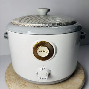 May include: A white NESCO electric cooker with a gray rim and lid. The cooker has a gold-colored dial with the brand name "NESCO" and a temperature control knob. The lid has a mesh screen. The cooker is on a light brown surface.