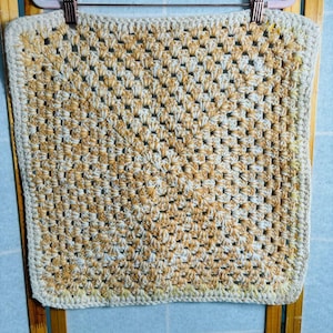 May include: A square, handmade crochet item with a granny square pattern. The yarn is a mix of cream, light blue, and golden yellow, creating a textured, warm appearance. The edges are finished with a cream-colored border.