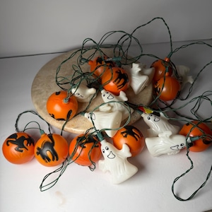 May include: A string of Halloween decorative lights featuring alternating orange plastic spheres with black cat silhouettes and white plastic ghost figures. The lights are connected by a dark green electrical cord and are arranged in a cluster.