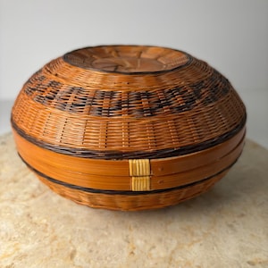 May include: A round, woven basket with a lid. The basket is made of brown and black wicker, with a decorative pattern around the top. The basket is sitting on a light-colored surface.