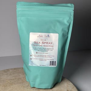 May include: A teal-colored bag of Dixie Belle Sea Spray texture additive. The label reads "Sea Spray - Texture Additive" and states it creates a chippy, textured, and weathered look. The bag contains enough Sea Spray for 2 quarts of Dixie Belle paint. It is 13 oz.