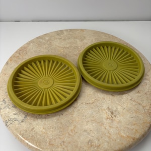 May include: Two olive green Tupperware lids with a sunburst design. The lids are circular with a raised center and ridged edges. They are displayed on a speckled, light brown surface.