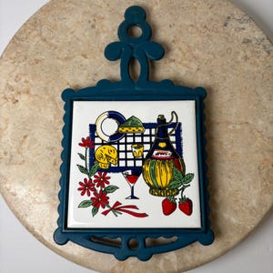 May include: A teal-colored trivet with a decorative border and handle. The center tile features a colorful illustration of a wine bottle, cheese, strawberries, and a glass of red wine. The trivet is designed to protect surfaces from hot items.