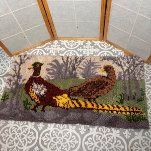 May include: A rectangular rug featuring two pheasants in a forest scene. The rug has a gray border and a green and brown background. The pheasants are brown, yellow, and green, with detailed feather patterns. The rug is likely handmade.