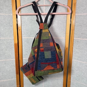 May include: A triangular-shaped backpack and a matching small pouch featuring a multi-colored woven tapestry pattern with geometric blocks of green, blue, orange, and red. The backpack has black adjustable straps and a vertical black zipper closure.