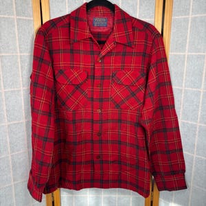 May include: A red plaid button-up shirt with black and yellow accents. The shirt has two chest pockets and a collar. The Pendleton label is visible at the top. The shirt is hanging on a black hanger.