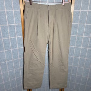 May include: Khaki-colored pleated pants hanging on a hanger. The pants have a button and zipper closure, with front pockets and a straight leg design. The fabric appears to be a durable cotton blend.