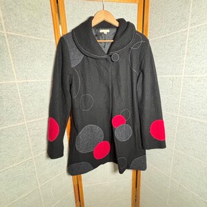May include: A black wool coat with a large collar and a single button closure. The coat features gray and red circular appliqués and white stitching details. The coat is hanging on a wooden hanger.