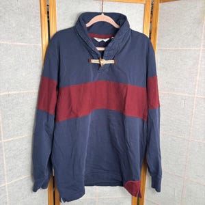 May include: A navy blue long-sleeve shirt with a burgundy stripe across the chest. The shirt has a shawl collar and a wooden toggle closure. The cuffs and collar are navy blue.