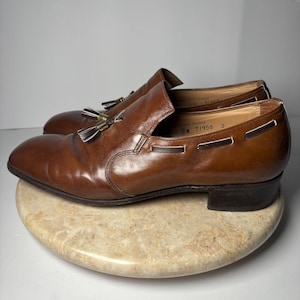 May include: A pair of brown leather loafers with tassel accents. The shoes have a classic design with a rounded toe and a low heel. The leather appears to be polished and in good condition. The shoes are displayed on a marble-like surface.
