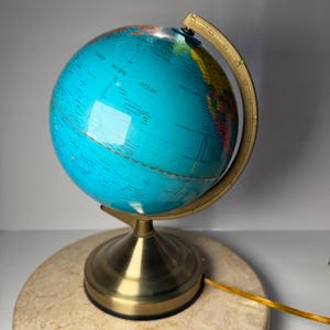 May include: A globe lamp featuring a bright blue ocean sphere with multicolored landmasses, mounted on a metallic gold-toned stand. The curved meridian arc includes printed degree markings, and a thin gold power cord extends from the base.