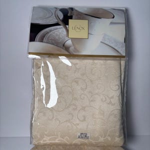 May include: A packaged ivory tablecloth with a subtle, repeating floral pattern. The packaging includes a Lenox logo and images of matching dinnerware. The label indicates the size fits a 60" x 102" oblong or oval table.
