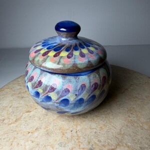 May include: A round ceramic jar with a matching lid, featuring a glossy finish and a repeating pattern of teardrop shapes in shades of blue, yellow, pink, and purple over a light blue and brown base. The lid has a small, rounded knob handle.