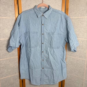 May include: Light blue short-sleeve button-down shirt with two chest pockets. The shirt is made of a denim-like material and hangs on a wooden hanger. The shirt has a classic collar and button closures.
