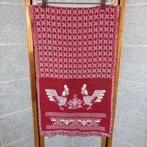 May include: A rectangular woven textile featuring a deep red background with white patterns. The upper section displays a repeating geometric star motif, while the lower section depicts two roosters flanking a central floral element.