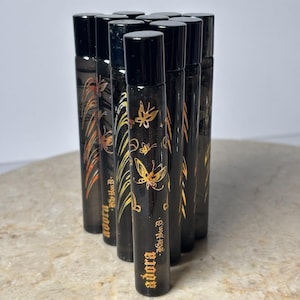 May include: A collection of black rollerball perfume bottles with black caps. The bottles are decorated with gold butterfly and floral designs, and the word "adora" is printed in gold on the front.