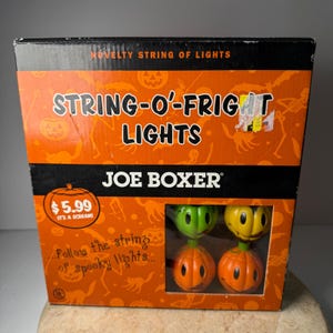May include: A rectangular cardboard box for Joe Boxer String-O-Fright novelty string of lights. The orange packaging features black text, pumpkin graphics, and a cutout window displaying four small, round, smiling pumpkin-shaped light covers.