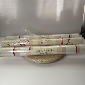 May include: Three rolls of Laura Ashley Home wallpaper. The wallpaper rolls are wrapped in clear plastic and feature a cream and burgundy plaid pattern. The brand name is printed on the label. The rolls are resting on a round, speckled, beige surface.