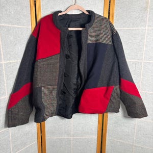 May include: A patchwork jacket with a round collar and black button closures. The jacket features panels of red, gray, and patterned fabric. The lining is black. The jacket is hanging on a wooden hanger.