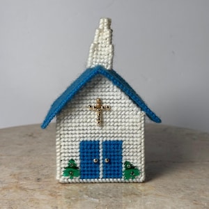 May include: A handcrafted miniature church with a white body, blue roof, and steeple. The church features blue double doors, a gold cross, and small green trees. The building is made of a textured material, possibly yarn or plastic canvas.