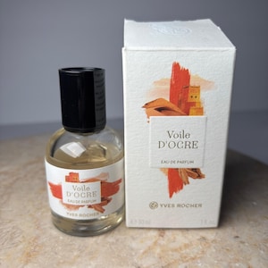 May include: A clear glass perfume bottle with a black cap and a white label featuring orange brushstroke graphics and the text Voile D'Ocre Eau de Parfum Yves Rocher, next to a matching white cardboard box with 30 ml and 1 fl. oz. printed on it.