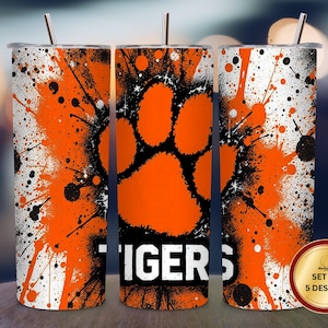 May include: A set of five stainless steel tumblers with a vibrant orange and black splatter design. Each tumbler features a large orange paw print and the word "TIGERS" in white. Includes a silver straw and a lid.