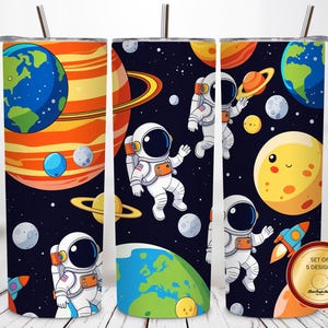 Może przedstawiać: Trzy cylindryczne kubki ze słomkami z motywem kosmicznym na ciemnym tle. Wzór zawiera rysunkowych astronautów, kolorowe planety, księżyce i rakiety. Złota, okrągła odznaka w rogu zawiera napis Set of 5 Designs.
