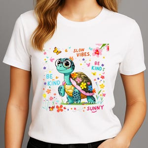 May include: White t-shirt featuring a cartoon turtle with a floral shell and the words "SLOW VIBES, BE KIND, SUNNY." The turtle is teal and yellow, surrounded by flowers and butterflies. A cheerful design.