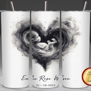 Könnte beinhalten: Weißer Becher mit einem schwarz-grauen Aquarelldesign eines Fötus in Herzform. Der Becher enthält den Namen "Emilia Rose W'son" und das Datum "21-10-2021". Enthält einen silbernen Strohhalm und ein Set mit 5 Designs.