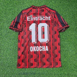 May include: Red and black Eintracht Frankfurt soccer jersey with the number 10 and the name OKOCHA on the back. The jersey has a geometric pattern and black accents on the sleeves. The word "Eintracht" is printed on the top.