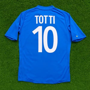 May include: A blue soccer jersey with the name "TOTTI" and the number "10" printed in white on the back. The jersey has short sleeves and a small gold star on the right sleeve.