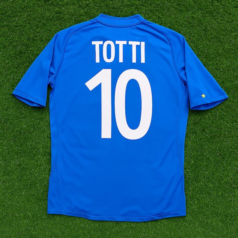 May include: A blue soccer jersey with the name "TOTTI" and the number "10" printed in white on the back. The jersey has short sleeves and a small gold star on the right sleeve.